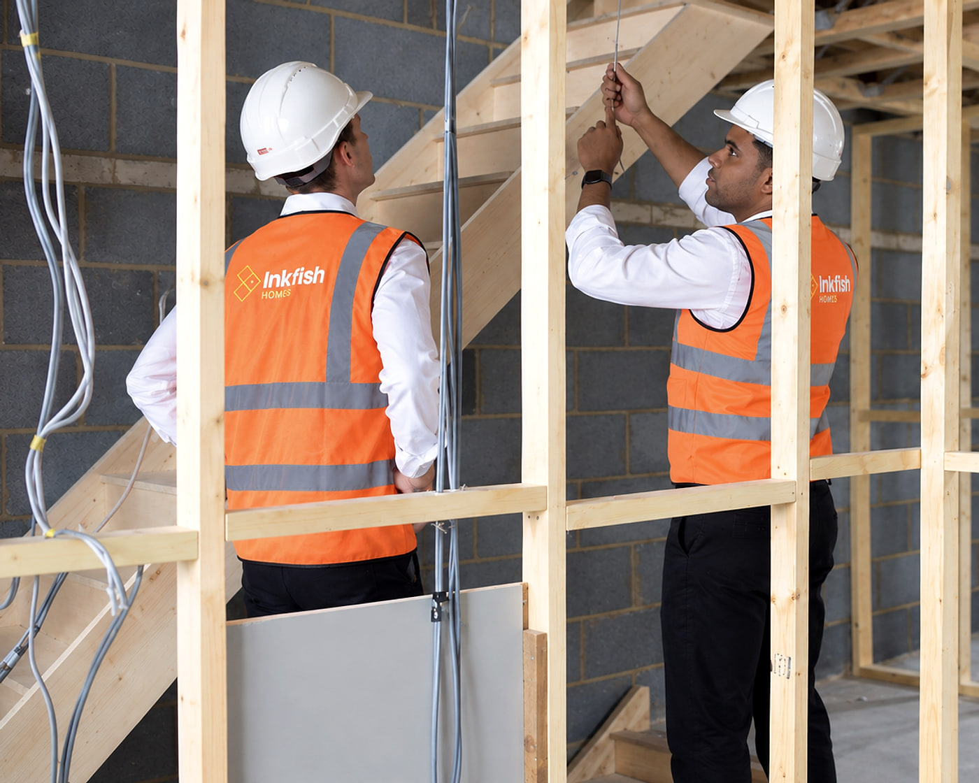 Louis Stedman-Bryce and colleague Elliot in Inkfish Homes high-visibility vests and hard hats, inspecting timber stud work inside a building under construction.